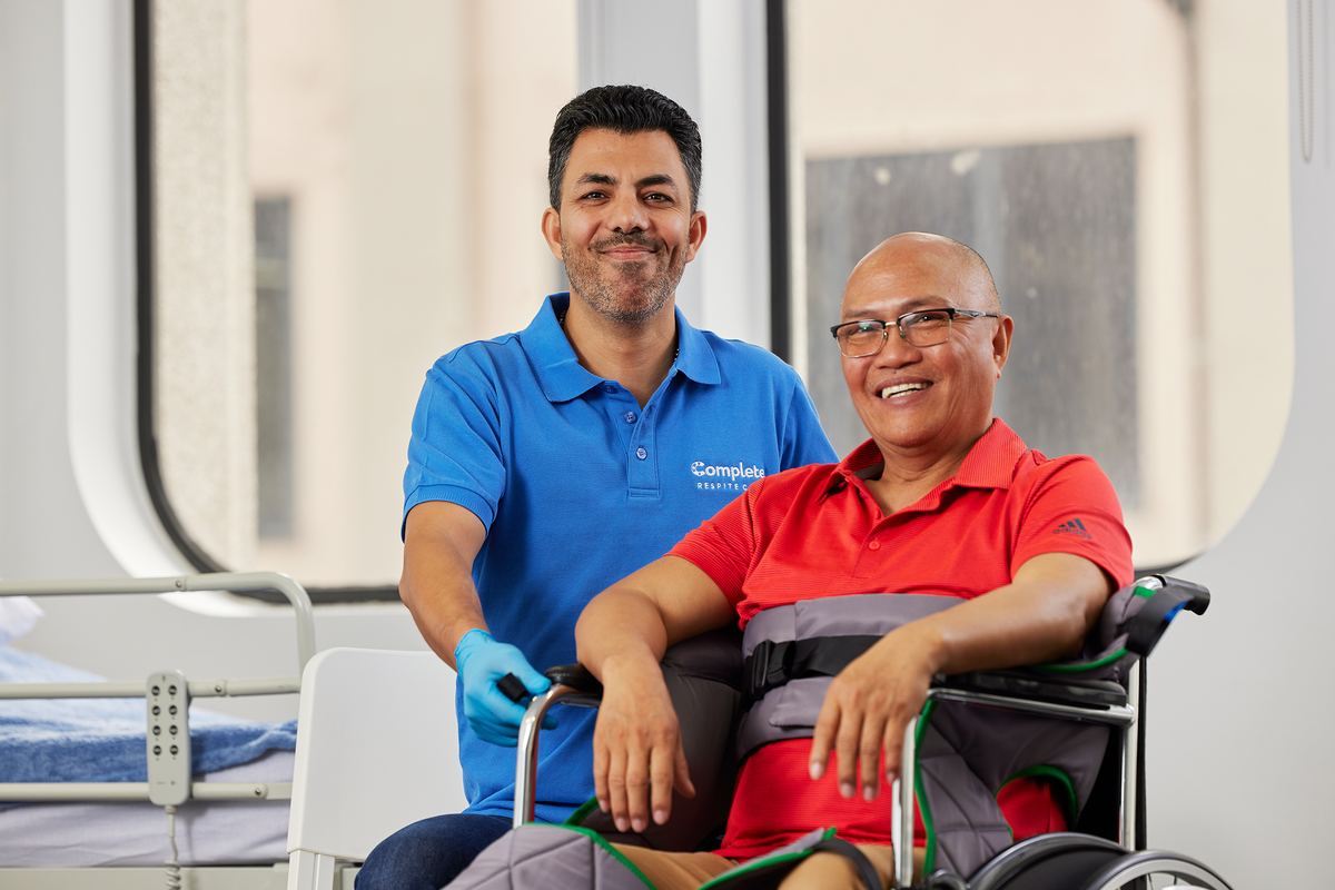 A carer standing proudly beside a seated older client, both smiling at the camera