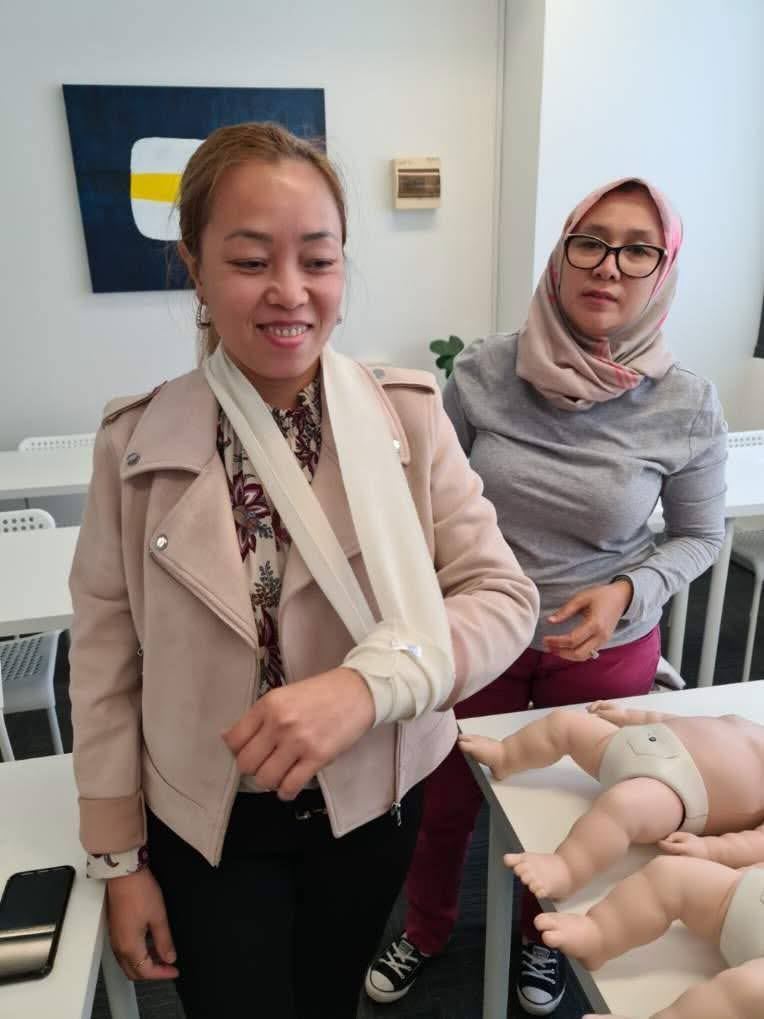 A trainee wearing a completed arm sling in a first aid class with infant CPR manikins on the table