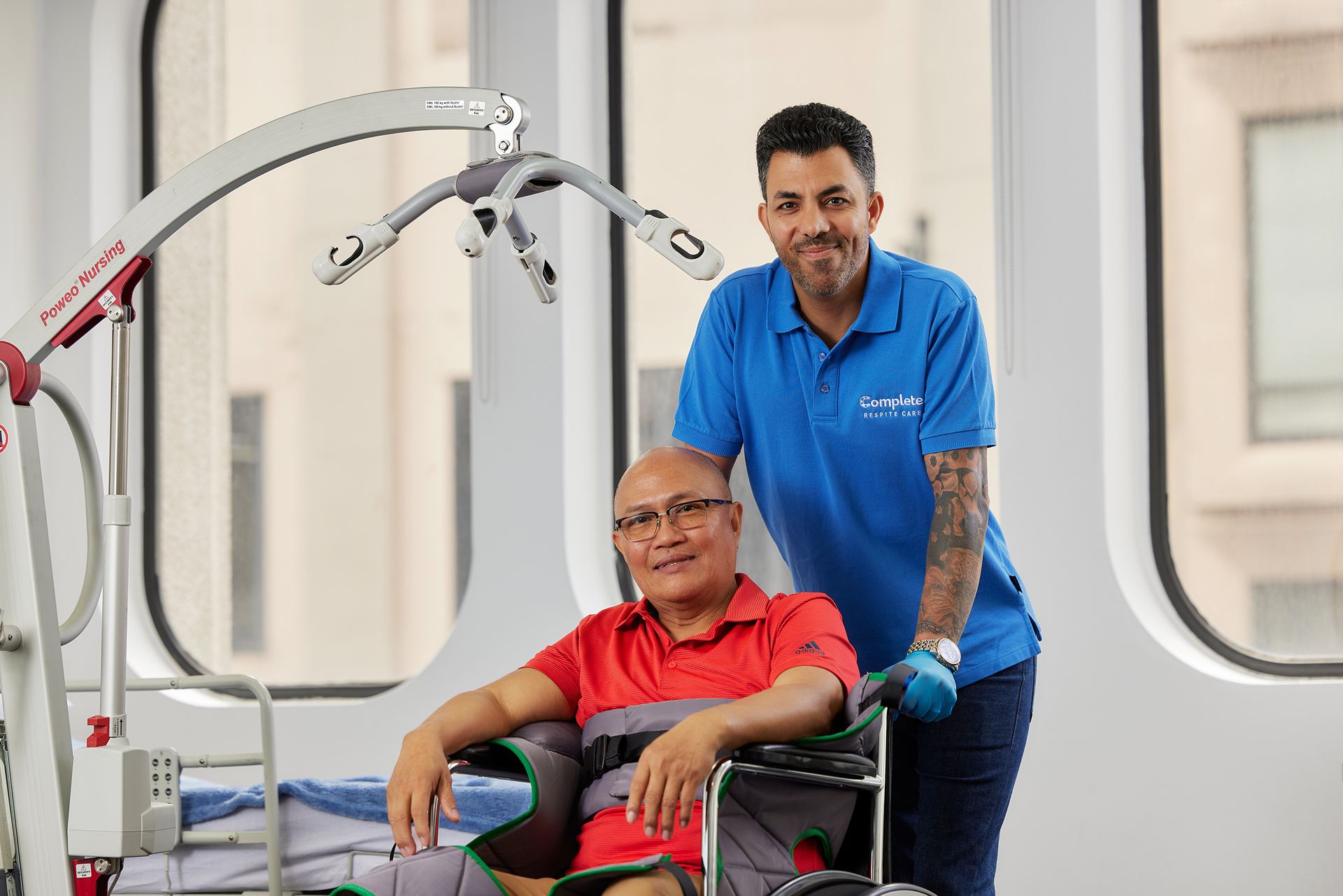 A smiling carer in uniform standing beside an older man seated in a wheelchair in a bright care facility