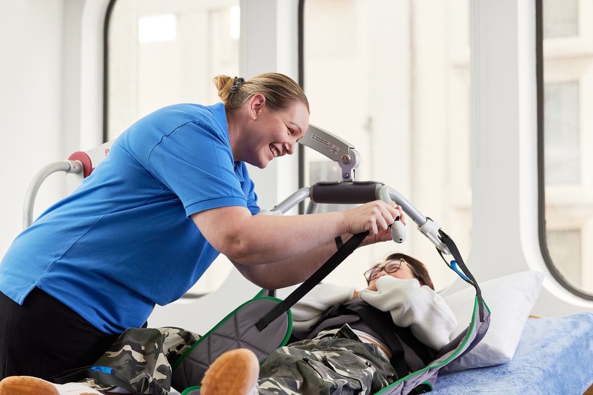 A trainer practising a patient hoist transfer with a client lying on a bed in a simulation lab