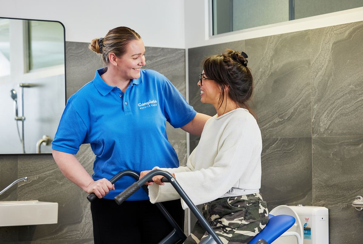 A carer assisting a young woman using a mobility aid in an accessible bathroom, both smiling